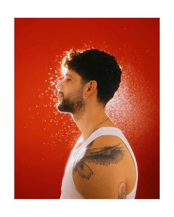 Tätowierter Mann im weißen Tanktop im Profil vor rotem Hintergrund, beleuchtet von Gegenlicht mit glitzernden Wassertropfen in der Luft
