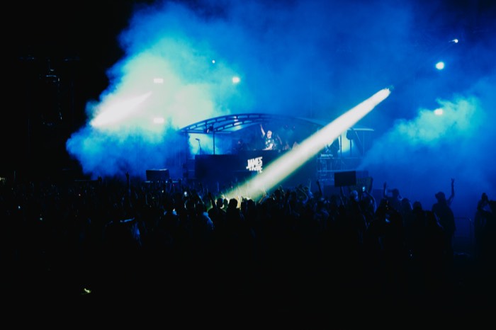 Ein DJ steht auf einer Festivalbühne im blauen Nebel, während ein greller Lichtstrahl über die silhouettierte Menschenmenge strahlt
