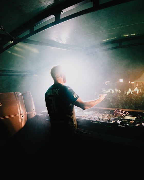 Ein DJ steht von hinten am Mischpult im hellen Gegenlicht und Nebel auf einer Festivalbühne vor dem Publikum