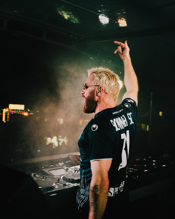 DJ James Hype steht mit erhobenem Arm am Mischpult einer dunklen Festivalbühne, beleuchtet von warmem Bühnenlicht im Nebel