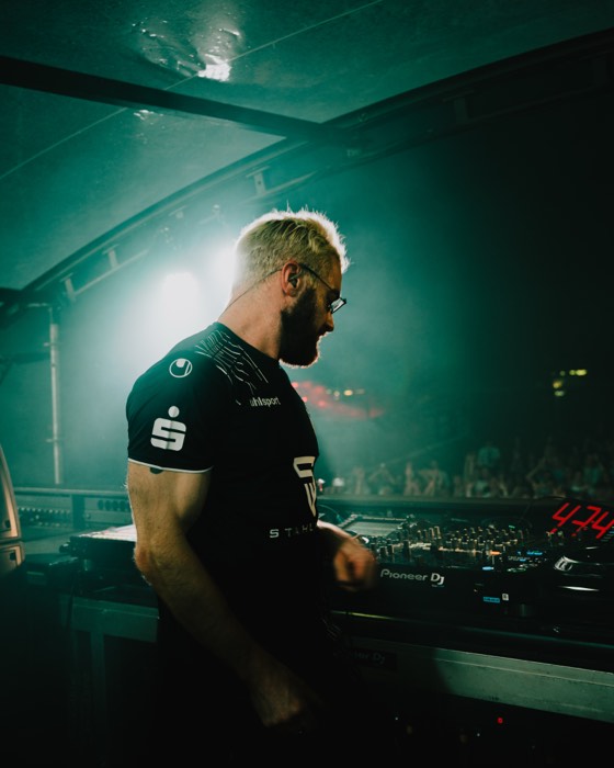 DJ James Hype steht im grünen Scheinwerferlicht und Nebel auf einer Festivalbühne an einem Mischpult