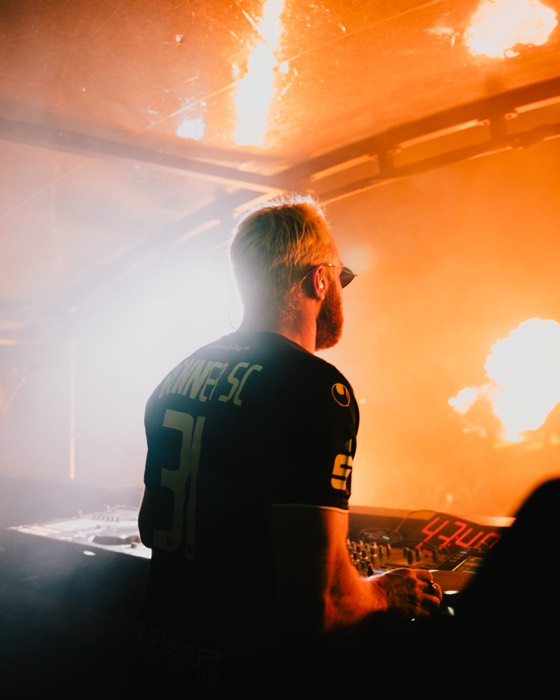 DJ James Hype steht von hinten am DJ-Pult auf einer Festivalbühne im hellen orangefarbenen Licht von Feuer und Nebel