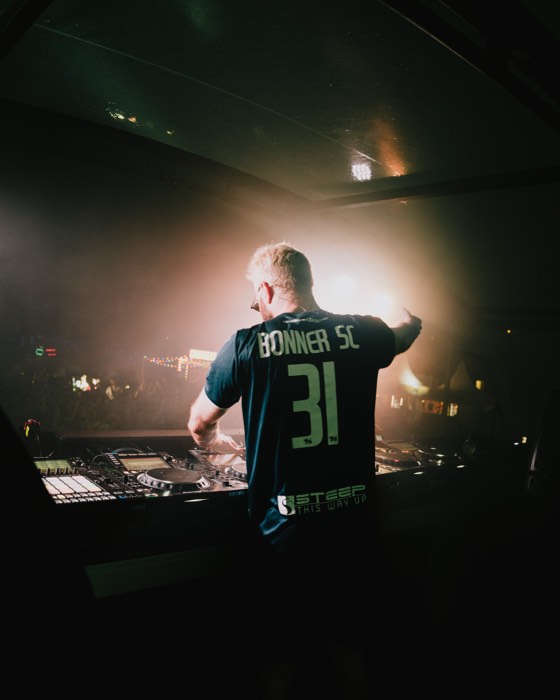 DJ James Hype steht von hinten am Mischpult einer Festivalbühne und gestikuliert im warmen Gegenlicht zum nächtlichen Publikum