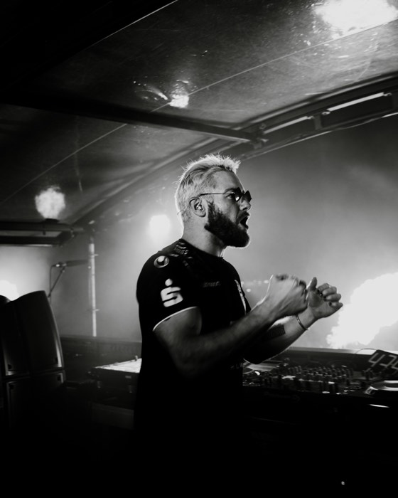James Hype jubelt mit geballten Fäusten am DJ-Pult einer vernebelten Festivalbühne bei starkem Gegenlicht und Pyrotechnik