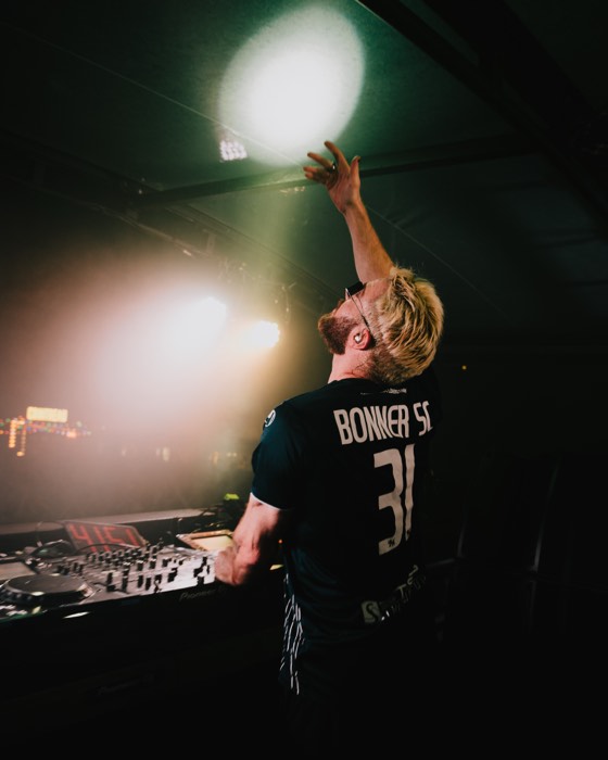 DJ James Hype steht im Trikot am Mischpult auf der dunklen Festivalbühne und streckt den Arm ins helle Scheinwerferlicht