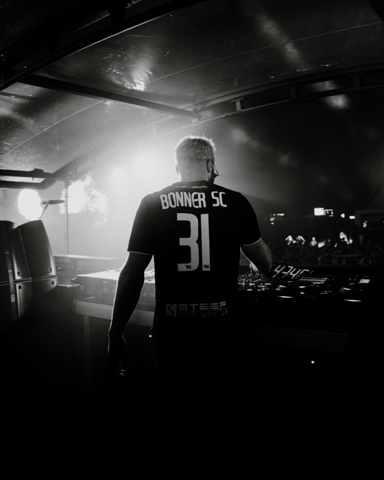Ein DJ in Rückansicht mit Bonner-SC-Trikot steht an seinem Pult auf einer Festivalbühne im hellen Gegenlicht der Scheinwerfer