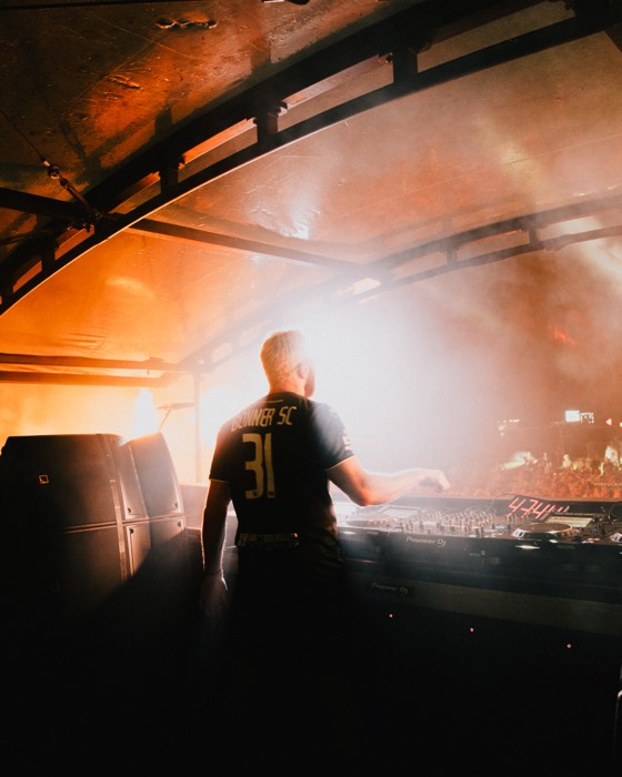 Ein DJ im schwarzen Trikot steht von hinten am Mischpult einer nebligen Festivalbühne im warmen, orangen Gegenlicht