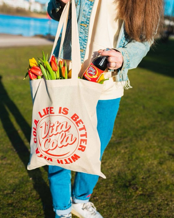 Eine Person hält im sonnigen Park einen bedruckten Vita-Cola-Stoffbeutel mit bunten Tulpen und greift nach einer Vita-Cola-Flasche