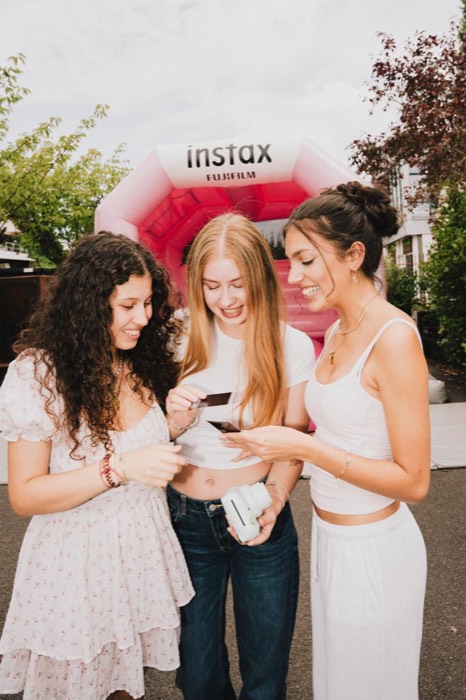 Drei junge Frauen mit Sofortbildkamera betrachten lächelnd ihre Fotos im hellen Tageslicht vor einem rosa Instax-Stand