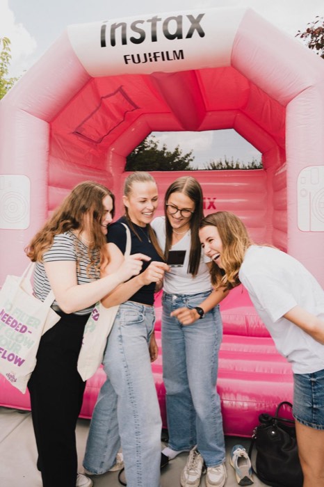 Vier junge Frauen betrachten lachend ein Sofortbild vor einer pinken Instax-Hüpfburg im hellen Tageslicht