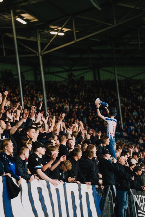 Fußballfans klatschen auf einer überdachten Tribüne im Stadionlicht, während ein Mann im gestreiften Trikot ein Megafon hochhält