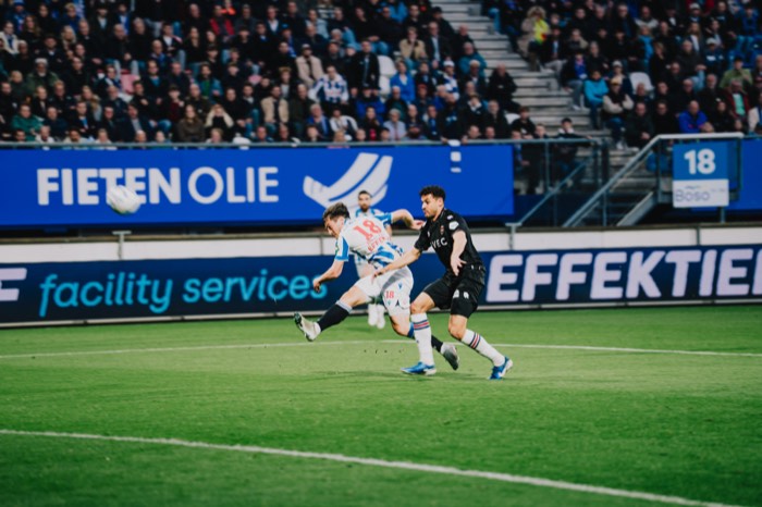 Ein Fußballspieler im blau-weißen Trikot schießt im hellen Stadion den Ball auf dem grünen Rasen an einem Gegenspieler vorbei