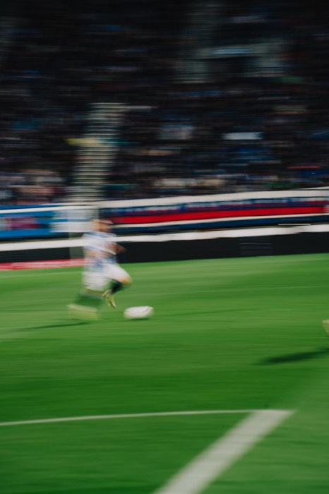 Ein Fußballspieler dribbelt bei Tageslicht in dynamischer Bewegung mit dem Ball über den grünen Rasen vor besetzten Tribünen