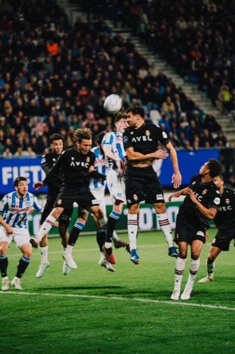 Fußballspieler in schwarzen und blau-weiß gestreiften Trikots springen unter Flutlicht im Stadion zum Kopfballduell hoch