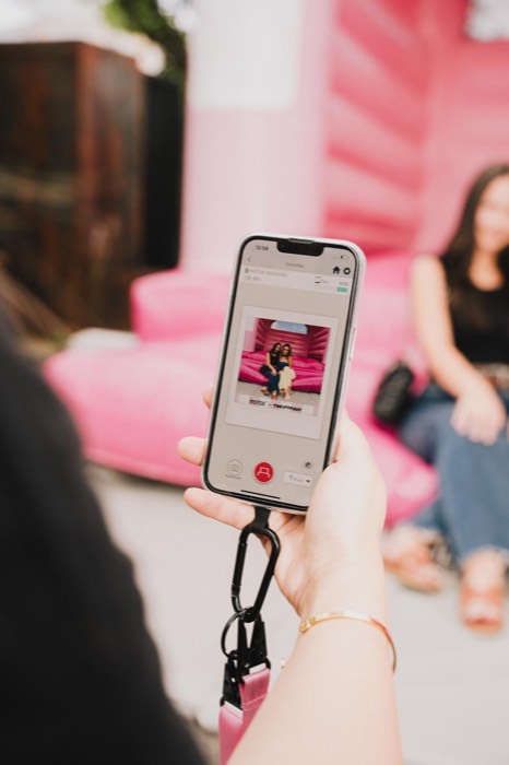 Eine Hand hält ein Smartphone mit einer Instax-App-Vorschau vor einem unscharfen pinken Hüpfburg-Hintergrund bei Tageslicht