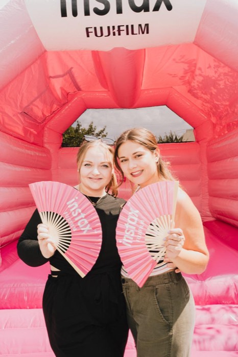 Zwei lächelnde Frauen stehen in einer pinken Hüpfburg und halten rosa Instax-Fächer bei hellem Tageslicht vor sich