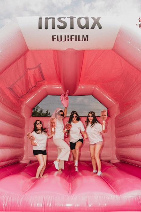 Vier Frauen springen bei hellem Tageslicht mit Foto-Requisiten in einer riesigen rosa Fujifilm-Instax-Hüpfburg
