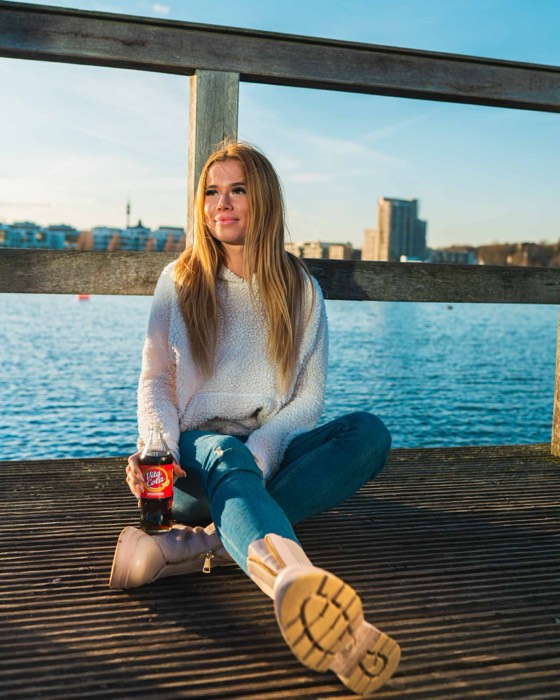 Eine Frau sitzt im warmen Abendlicht auf einem Holzsteg an einem See und hält eine Flasche Vita Cola in der Hand