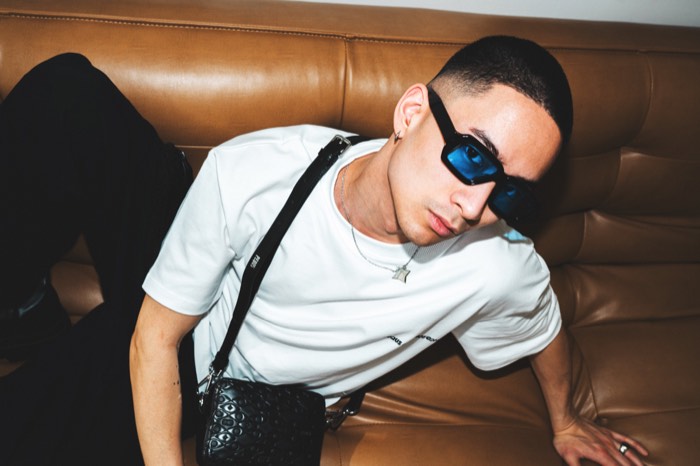 Ein Mann mit blauer Sonnenbrille und weißem T-Shirt lehnt mit Schultertasche auf einer braunen Ledercouch bei direktem Blitzlicht