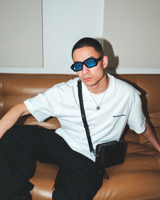 Mann mit blauer Sonnenbrille, weißem Shirt und schwarzer Tasche sitzt auf einer braunen Ledercouch bei direktem Blitzlicht