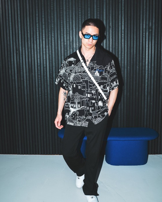 Ein Mann im schwarz-weiß bedruckten Hemd und blauer Sonnenbrille steht vor einer dunklen Lamellenwand bei hartem Blitzlicht