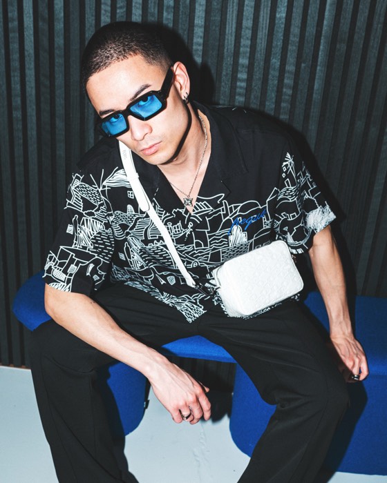 Mann mit blauer Sonnenbrille und weißer Tasche sitzt im harten Blitzlicht auf einer blauen Bank vor dunkler Lamellenwand