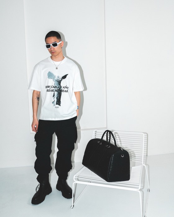 Ein Model in weißem Grafik-Shirt und Sonnenbrille steht im hell ausgeleuchteten Studio neben einer Tasche auf einem Stuhl