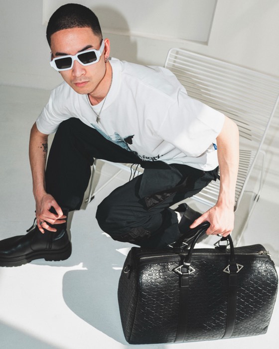 Ein Mann mit weißer Sonnenbrille hockt bei hartem Licht im hellen Studio und hält eine schwarze Reisetasche