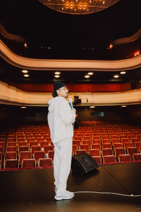 Tony Bauer steht in weißer Kleidung mit Mikrofon auf einer Bühne im warm erleuchteten, leeren Theatersaal