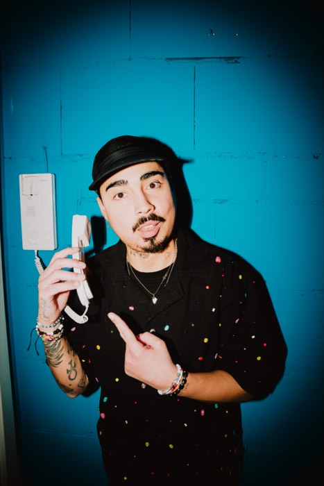 Tony Bauer mit Cappy zeigt im harten Blitzlicht vor einer blauen Wand auf den weißen Telefonhörer in seiner Hand