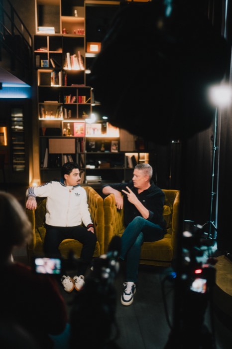 Tony Bauer und ein Mann unterhalten sich vor Kameras auf gelben Sesseln in einem warm beleuchteten Studio