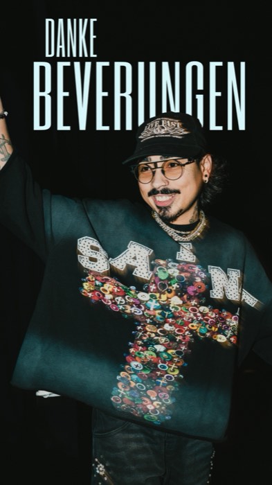 Tony Bauer lächelt mit Cap und Brille mit erhobenem Arm vor schwarzem Hintergrund im hellen Blitzlicht