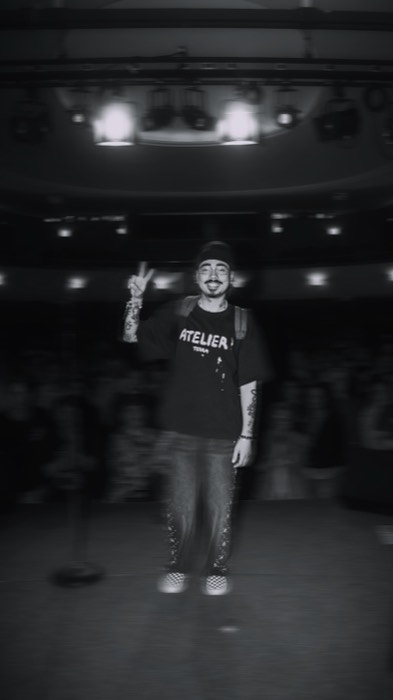 Tony Bauer macht lächelnd das Peace-Zeichen auf einer dunklen Bühne vor Publikum, beleuchtet von hellen Scheinwerfern