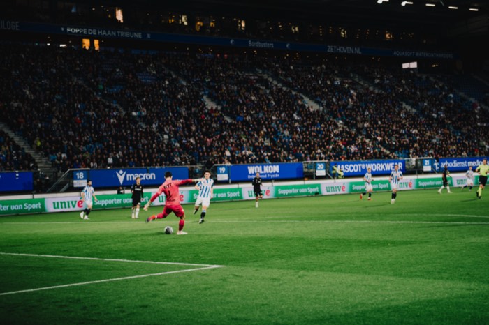 Ein Torwart im roten Trikot schießt den Ball auf dem grünen Rasen eines voll besetzten Fußballstadions unter Flutlicht