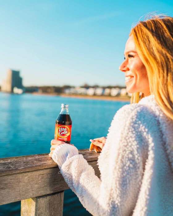 Eine lächelnde blonde Frau hält an einem Geländer am Wasser eine Vita Cola Flasche im warmen Sonnenlicht