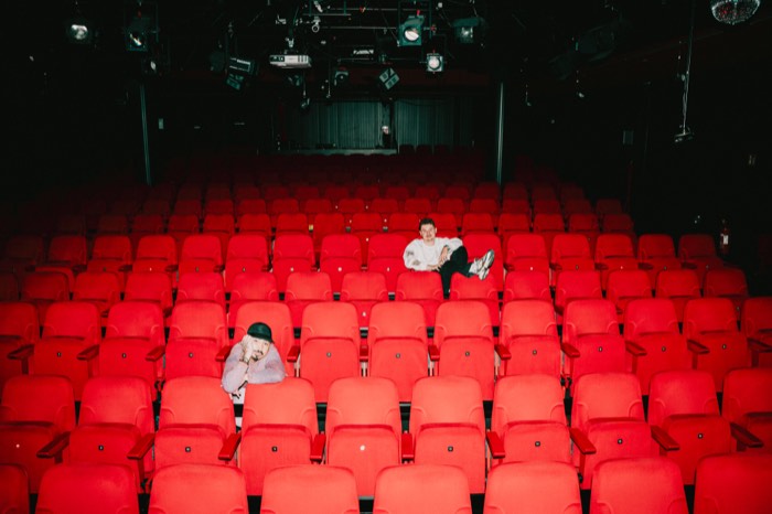 Zwei Männer sitzen verteilt in den roten Stuhlreihen eines leeren Theatersaals unter künstlichem Licht